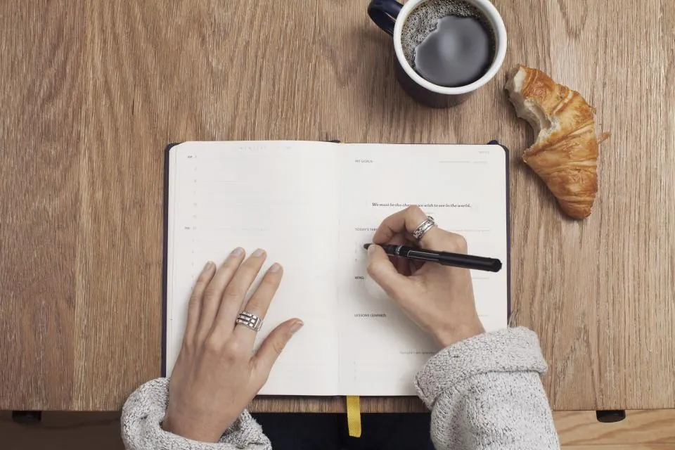 Hands writing in an open notebook on a wooden desk, next to a cup of coffee and a croissant.
