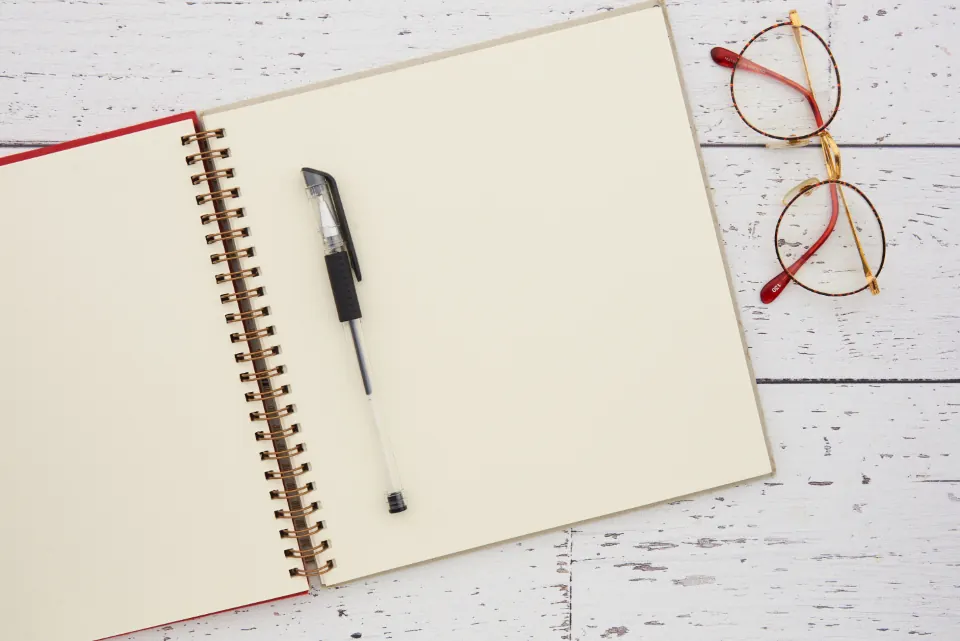An open spiral notebook and a pen on a white wooden table, next to a pair of glasses.