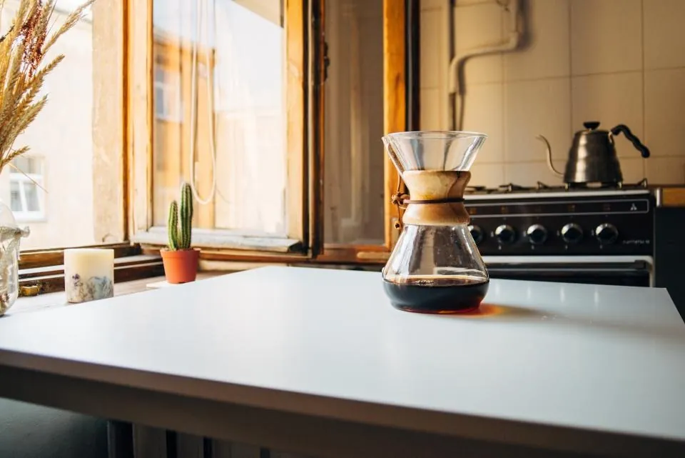 A white kitchen counter with a glass coffee maker, a kettle and a plant by a window.