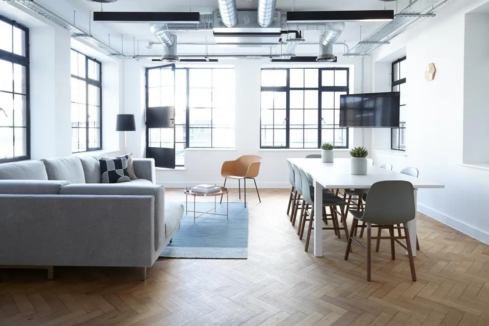 A bright open room with a grey sofa, a rug and a long dining table with chairs under tall windows.