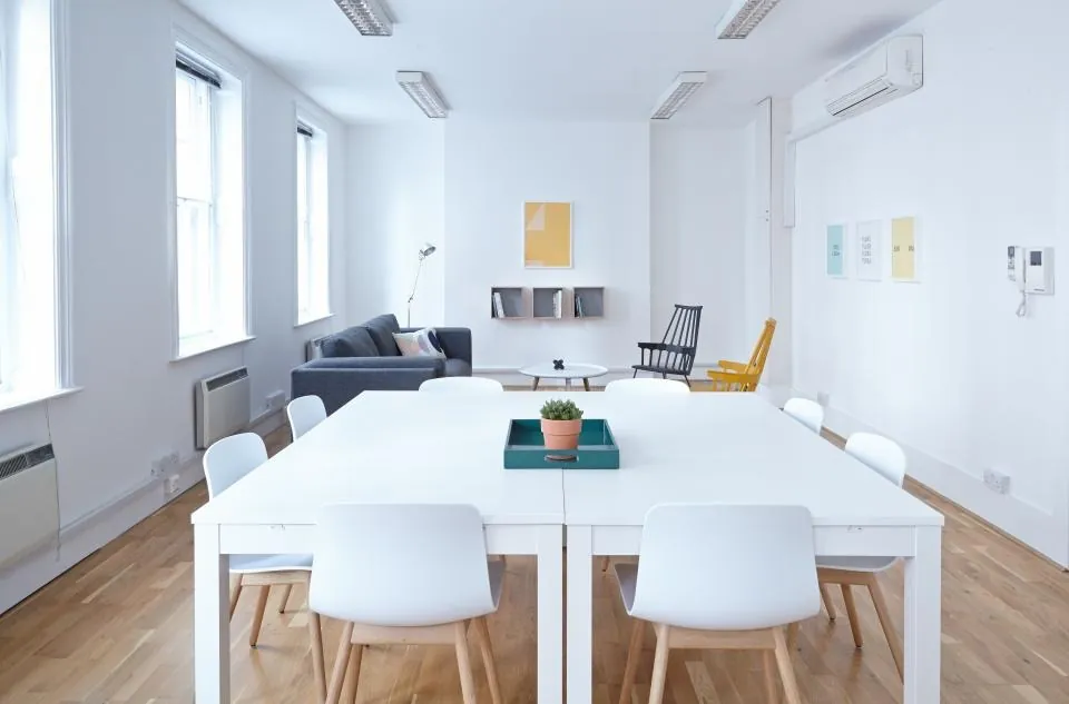 A white room with long shared tables, white chairs and a sofa at the far end.
