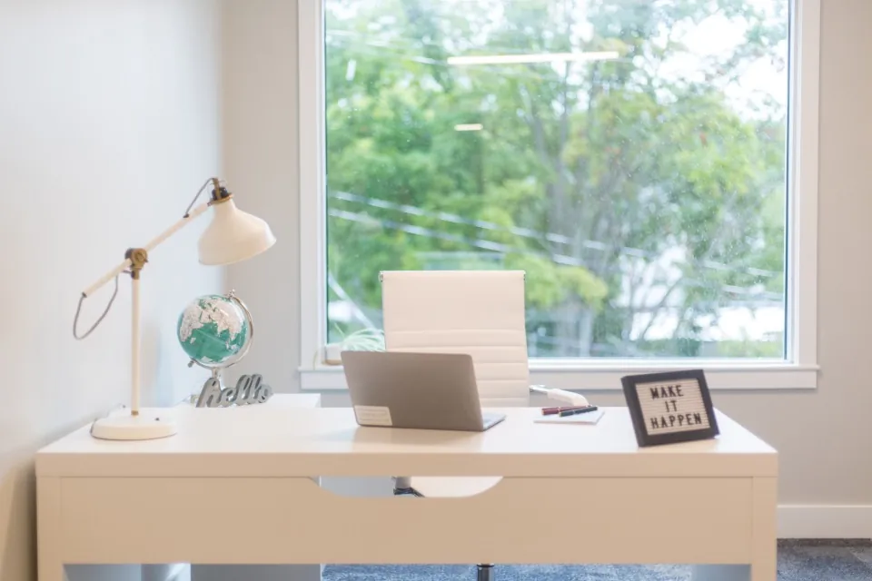 A white desk with a lamp and a closed laptop in front of a large window onto trees.