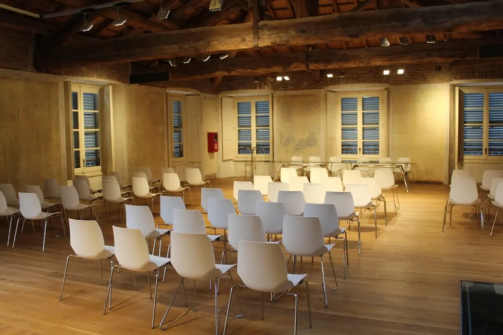 A bright hall with wooden ceiling beams, tall windows and rows of white chairs facing the front.