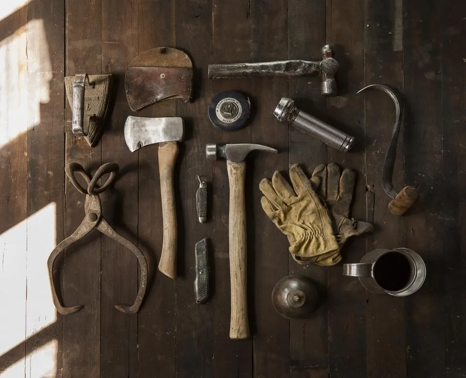 Hand tools laid out on a wooden bench: a hammer, pliers, a small axe, gloves and a few smaller tools.