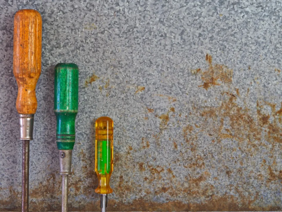 Three old screwdrivers with orange, green and yellow handles against a rusty grey metal sheet.