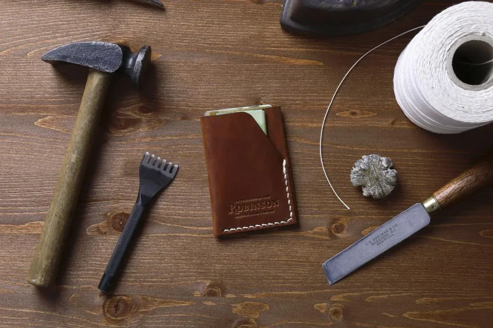 A hammer, a small chisel, a leather wallet, a cutting knife and a spool of thread on a dark wooden table.