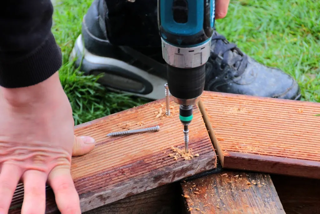 A close-up of hands drilling into a wooden deck board outdoors.