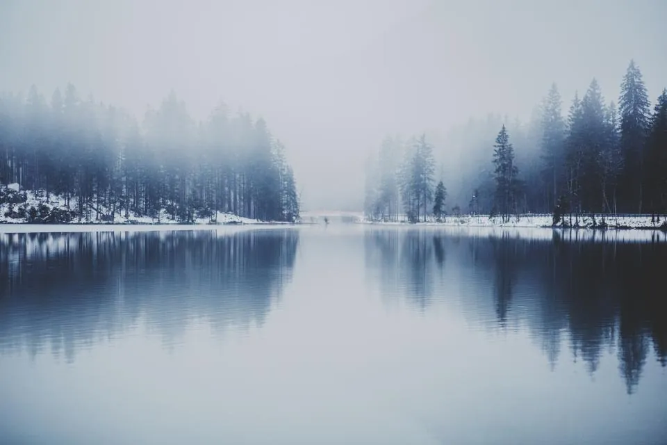 A still lake reflecting snowy pine trees and low grey cloud.