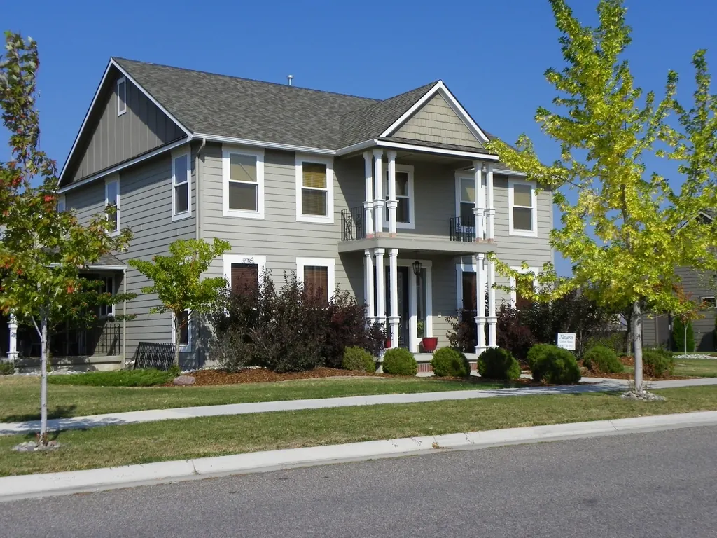 A grey two-storey house with a front porch and young trees on the pavement.