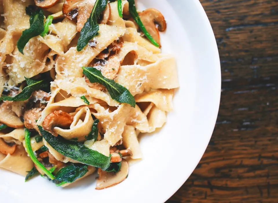 Wide ribbons of pasta with mushrooms and greens on a white plate.