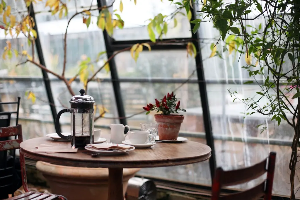 A round café table by a tall window with a cafetière, cups and potted plants.